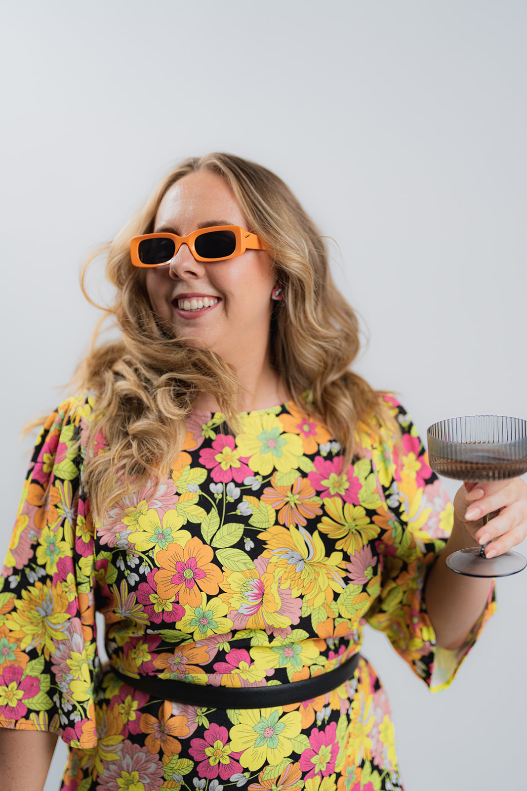 Female in a yellow floral dress with orange sunglasses holding a black cocktail glass. She is swishing her hair and looking to the left of the camera