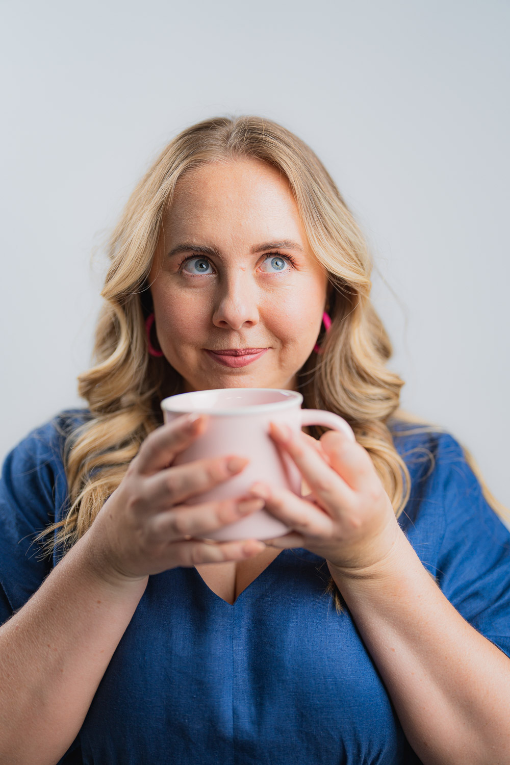 Girl in a blue dress with blonde hair and blue eyes is holding a coffee cup up to her face and is looking away from the camera